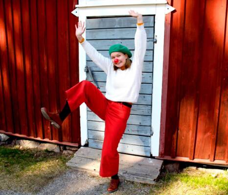 Punanenäinen klovni hymyilee ja nostaa käsiä ja jalkaa ilmaan. Takana on vanhan punamultaisen piharakennuksen suljettu ovi.