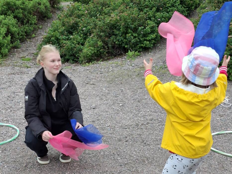Miisa kyykyssä maassa ja edessä on lapsi heittämässä huivia ilmaan.